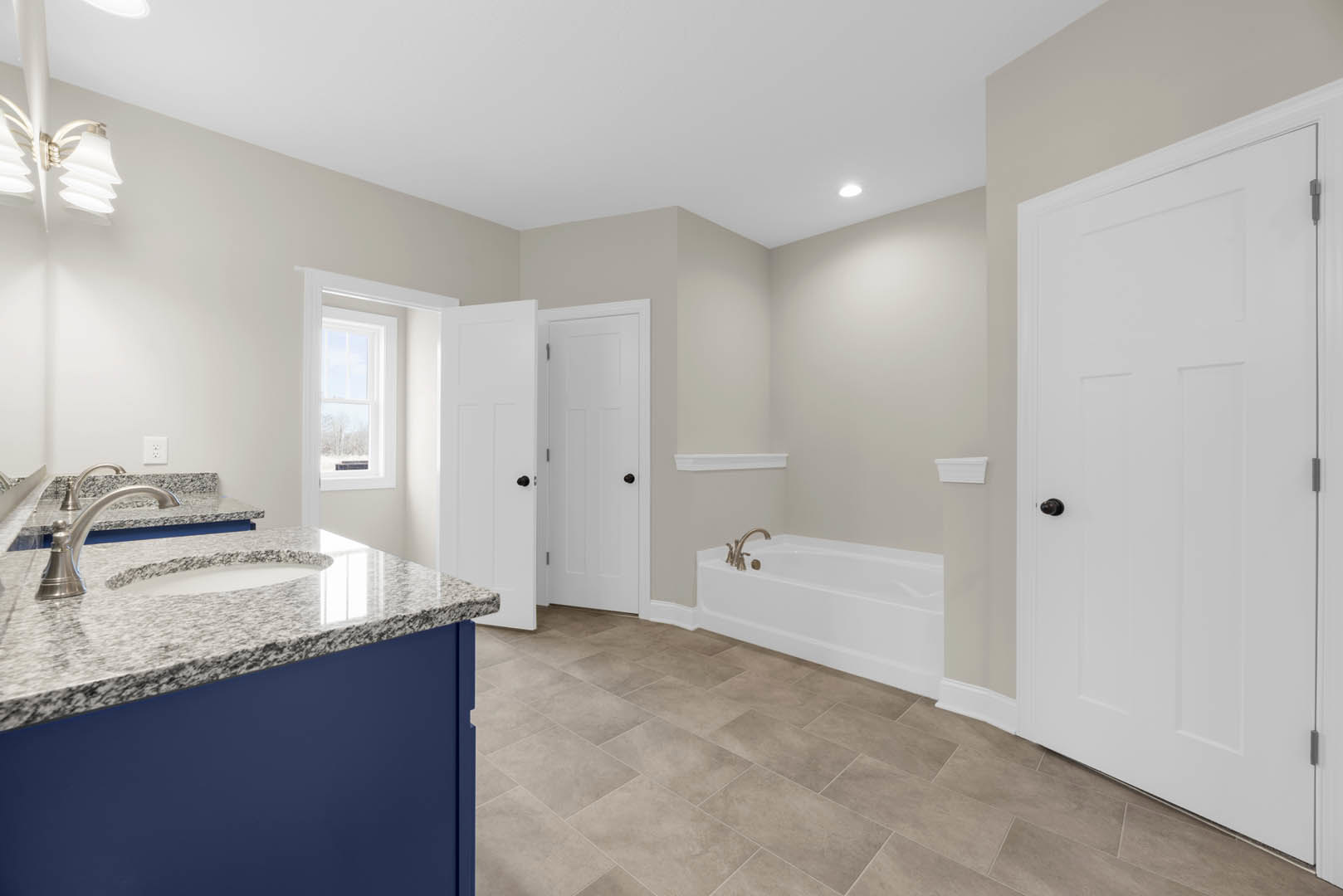 Bathroom with white tile floor, freestanding white bathtub with silver faucets, white vanity sink, black hardware on white door, and modern light fixture
