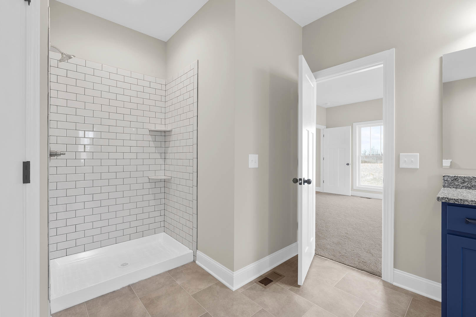 White tile shower with built-in shelf and white shower base, open door revealing carpeted floor, window offering outdoor view, white light switch on wall