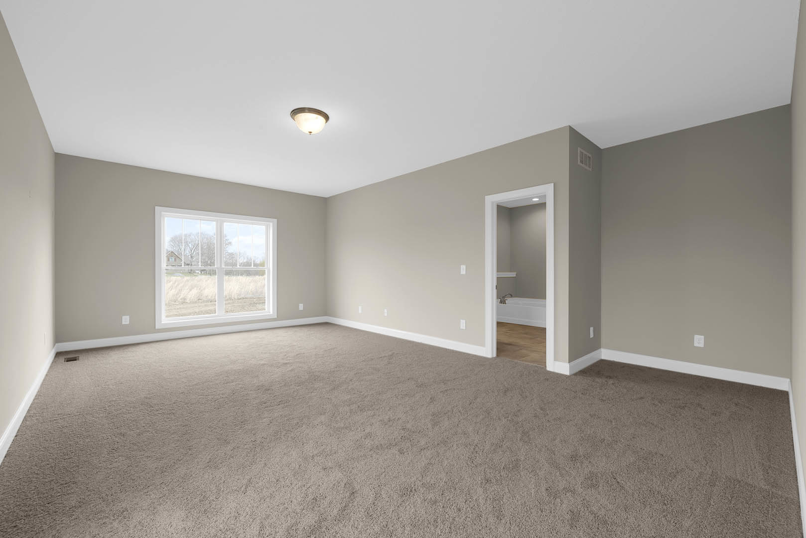 Bedroom with beige carpet, open white door, large window overlooking grassy field, ceiling light fixture, adjacent bathroom featuring a white bathtub and built-in shelf