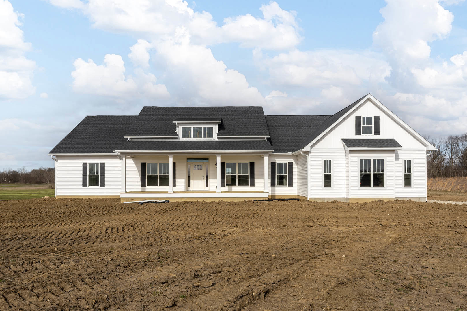 White farmhouse with black roof, multiple windows, and white door, surrounded by a dirt field with visible tire tracks under a cloudy sky.