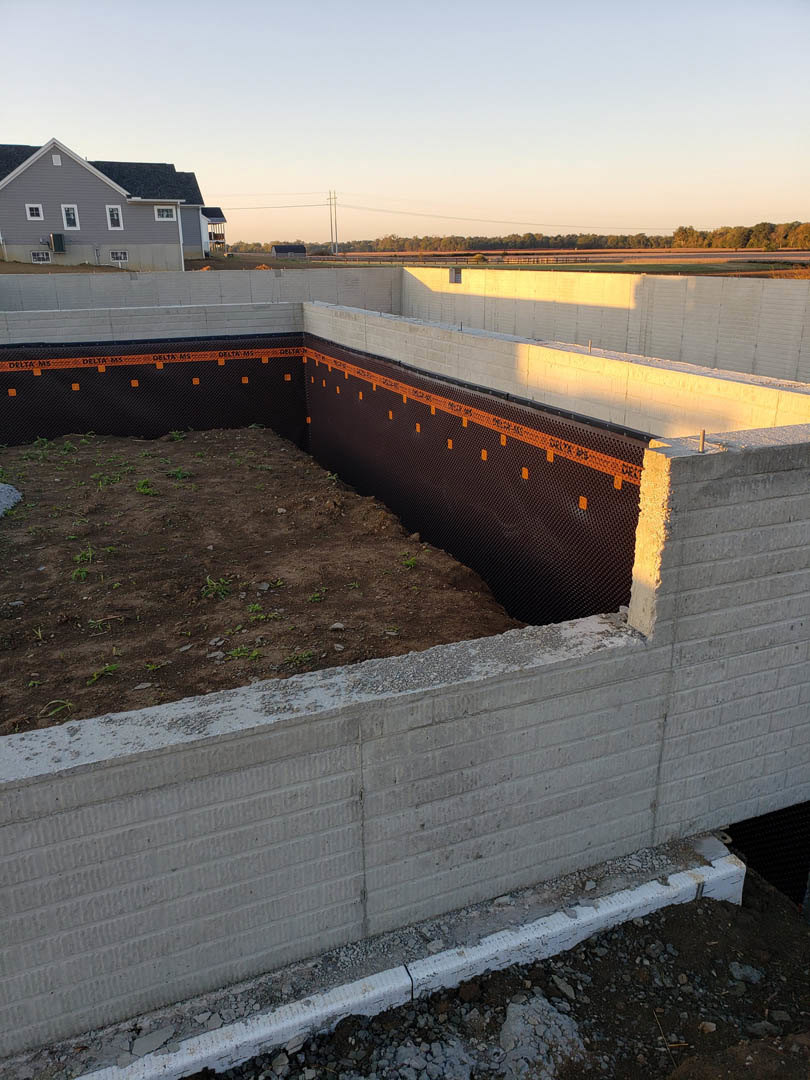 Partially built house with exposed foundation, white curb, large wall opening, and multiple windows, set against blue sky and surrounding trees.