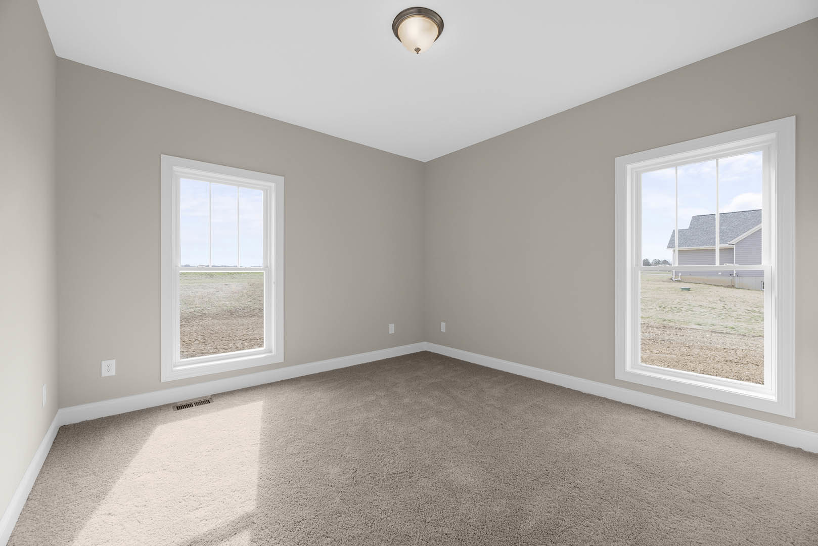 Carpeted room with two windows, white walls, and decorative molding; one window offers a view of a neighboring house, the other overlooks a grassy field; close-up of a modern light