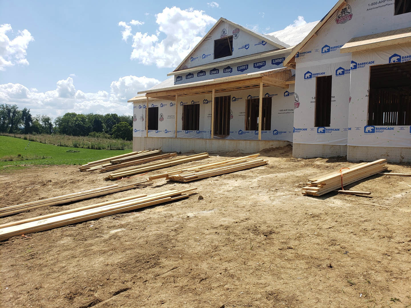 Framed house under construction with exposed wood planks, unfinished windows, and stacks of lumber on bare ground