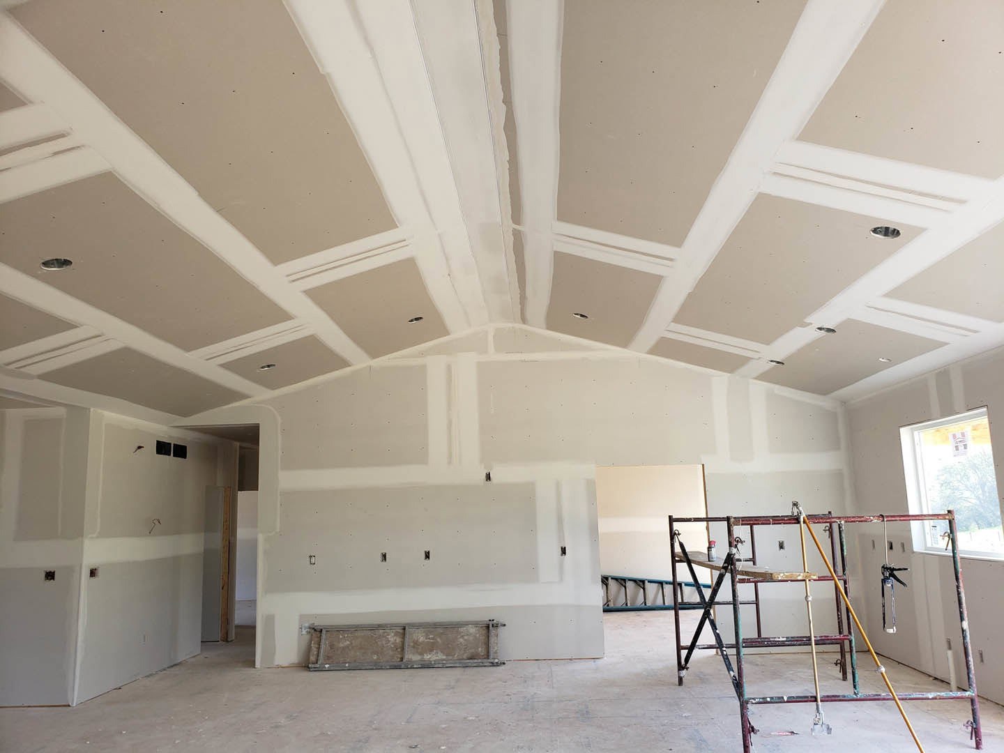 Scaffolding and ladder set up in a partially finished room with exposed wooden framing, white plaster surfaces, spray gun and painting tools, window with taped paper, and wall