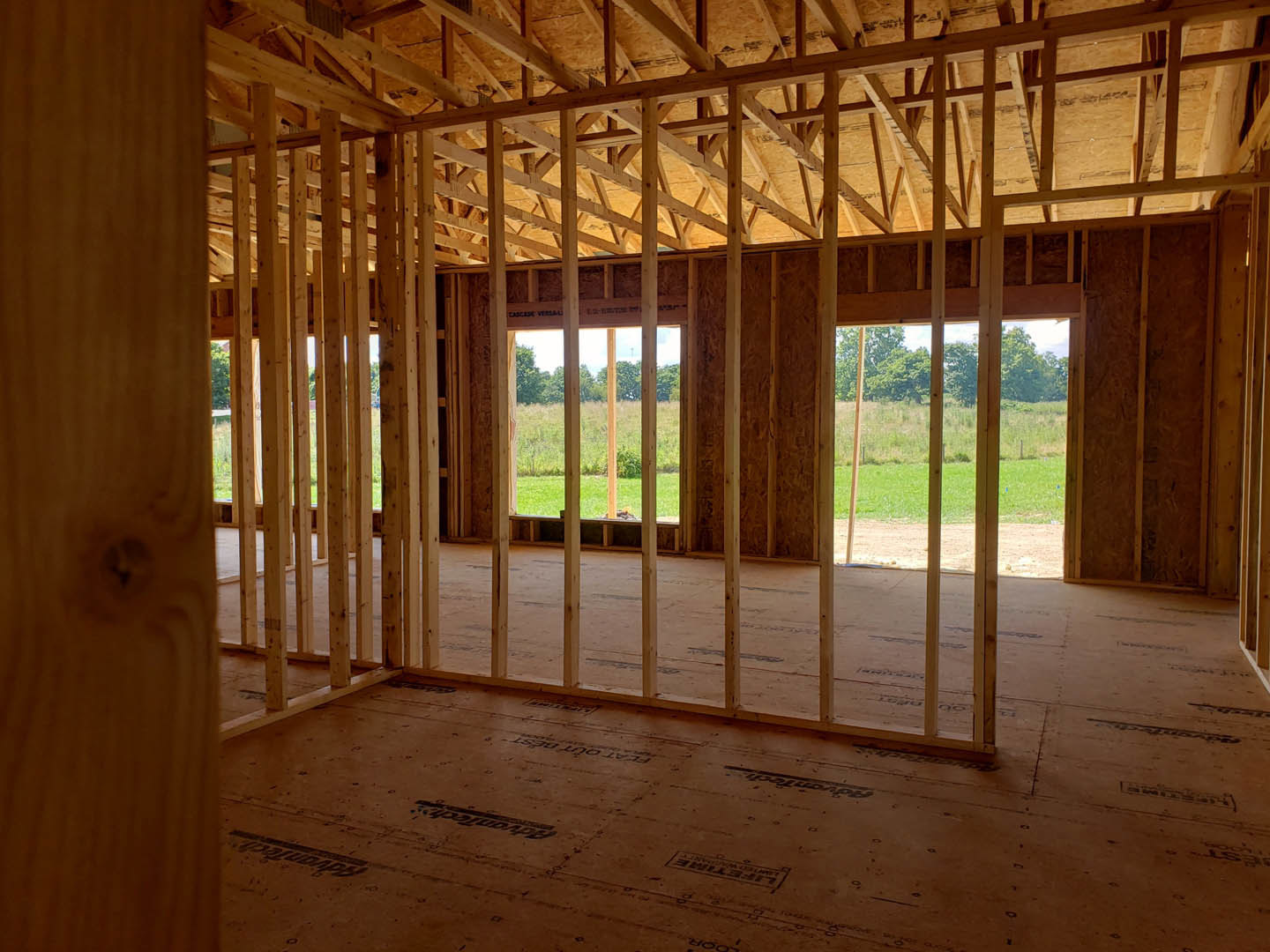 Wood-framed house under construction with exposed beams, unfinished floors, and large windows overlooking grassy field and trees