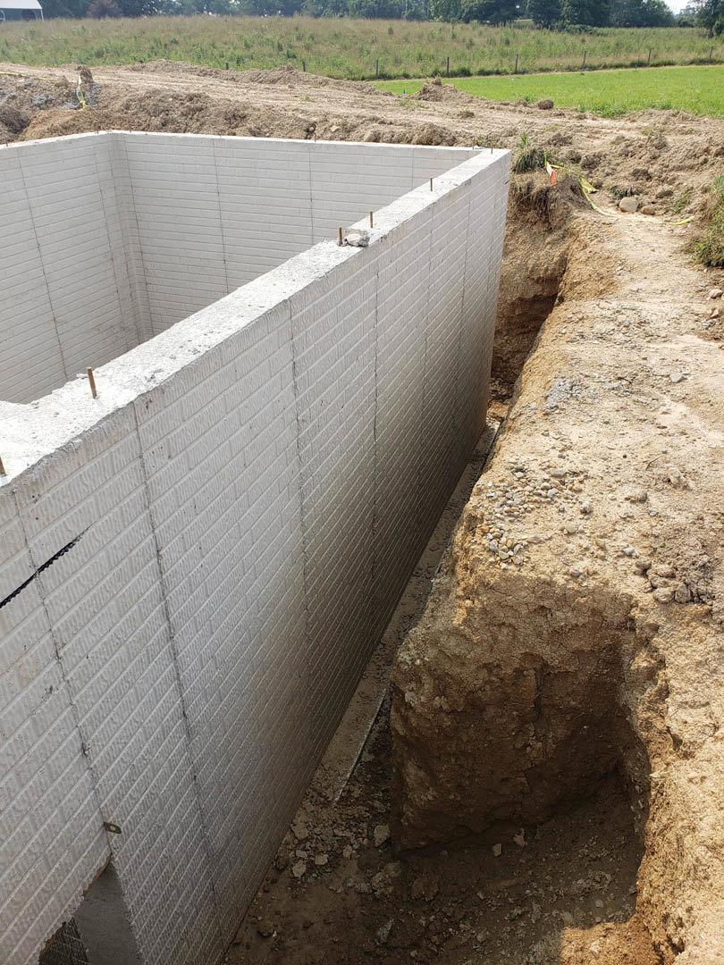 Concrete foundation wall with exposed dirt pit in foreground, surrounded by grassy field and trees.