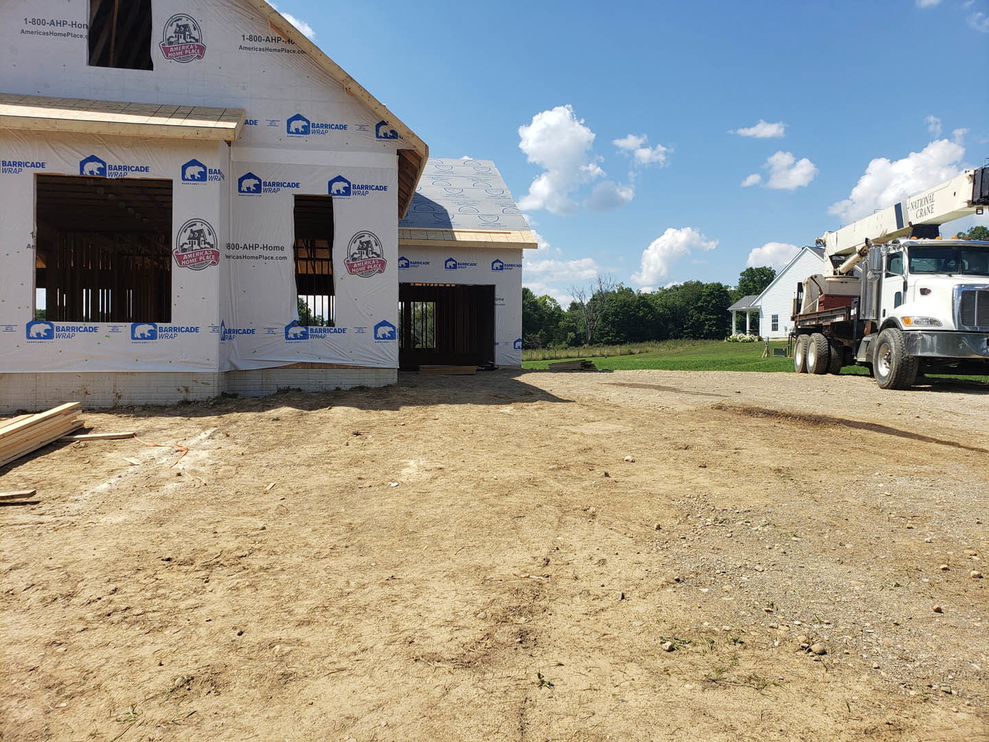 White plastic-wrapped house under construction on a dirt lot, crane truck parked in back, trees and cloudy sky in background