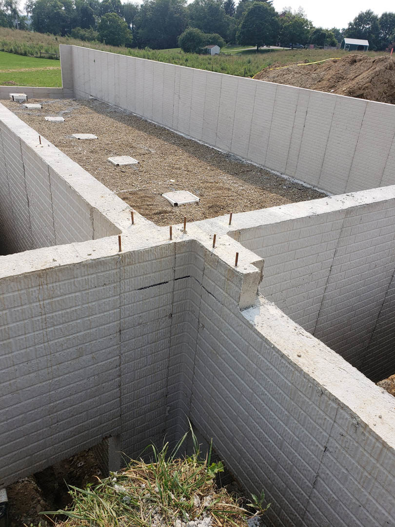 Concrete foundation with several square holes, surrounded by grass, rocks, and a pile of dirt with a yellow hose; group of trees in the background under a clear sky.