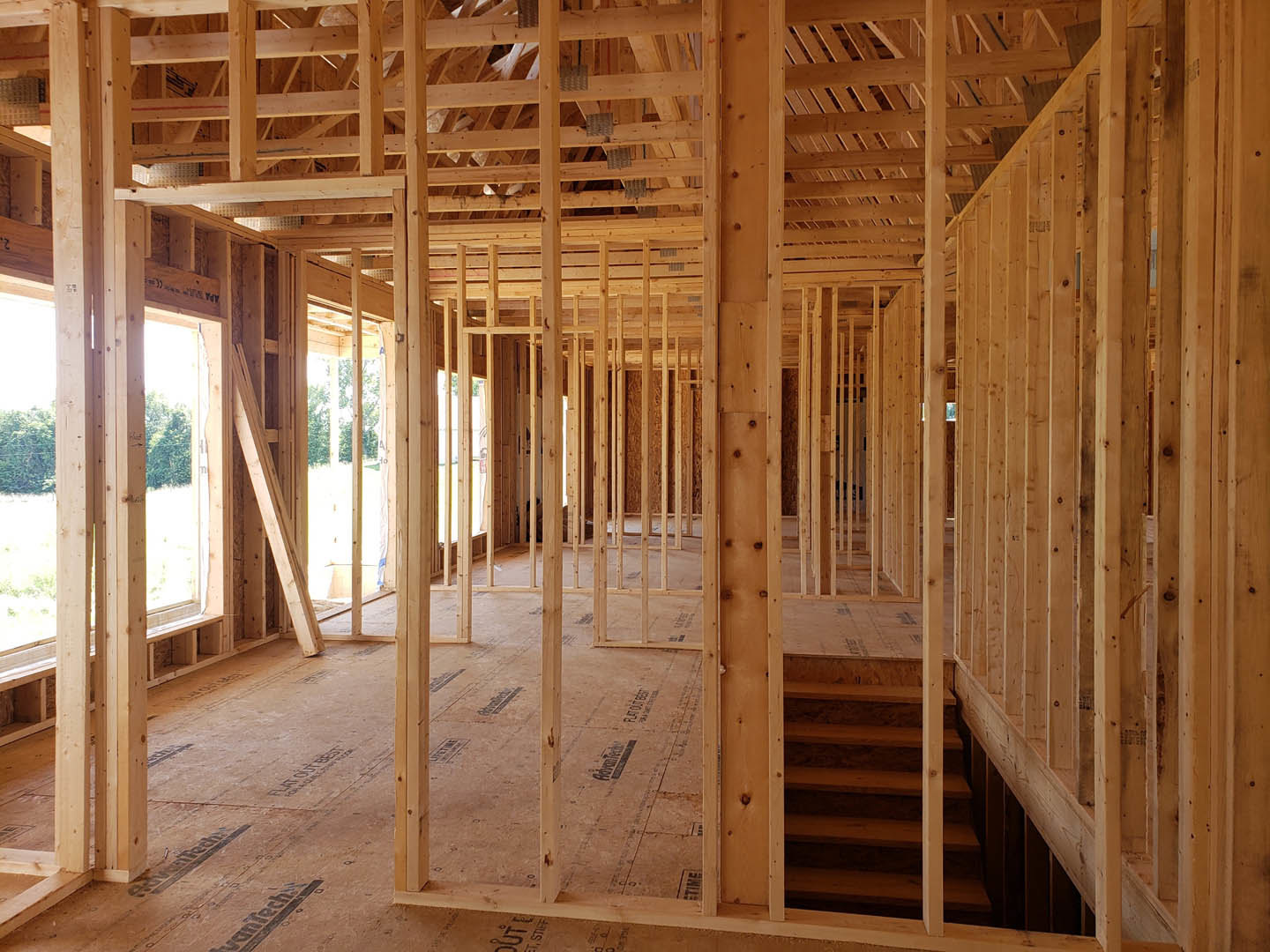 Exposed wooden framing and beams inside a house under construction, unfinished floors and ceiling, visible insulation between studs