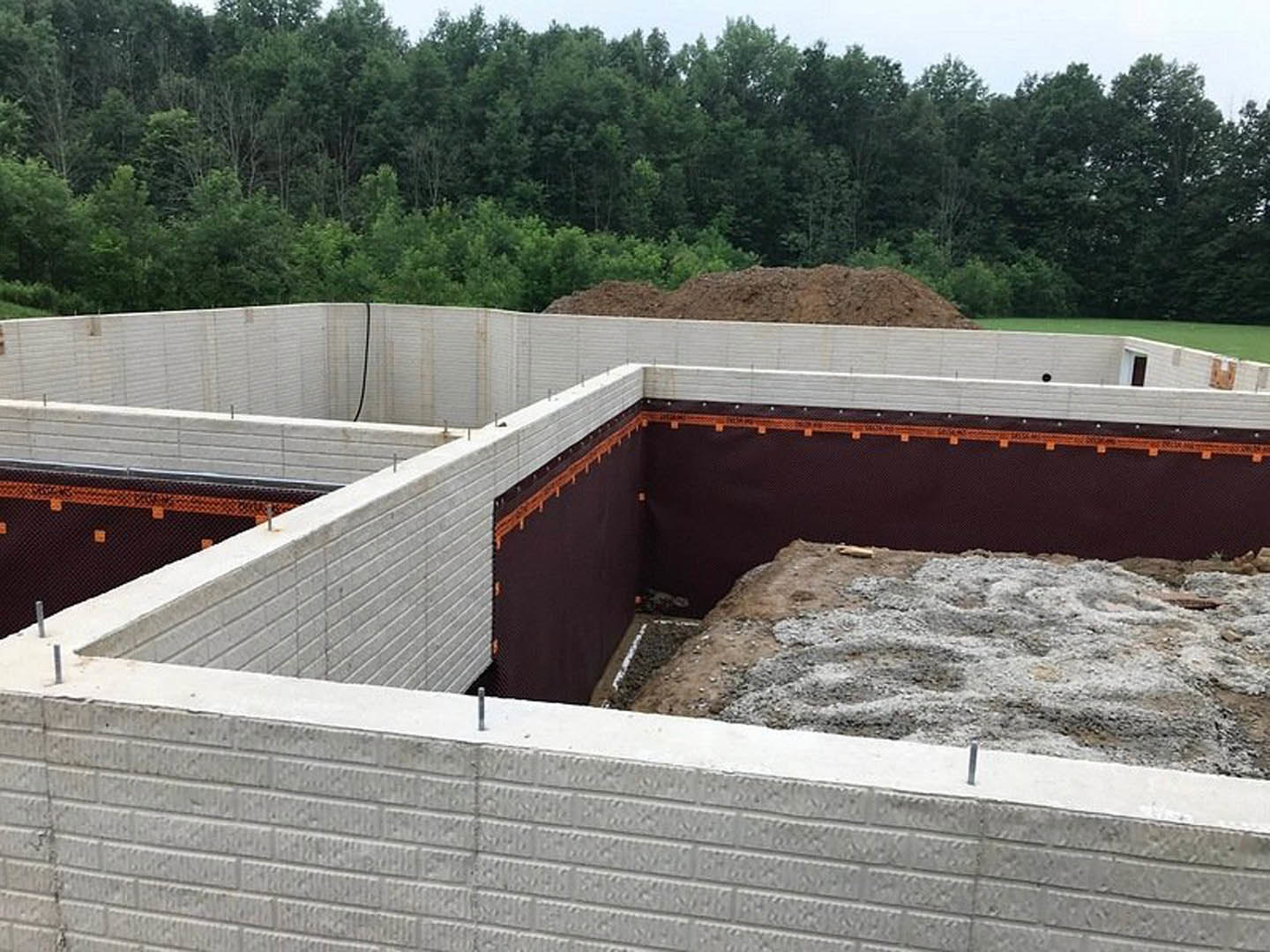 Concrete foundation slab with white brick perimeter wall, metal rebar protruding, piles of dirt along the edge, and trees in the background under open sky