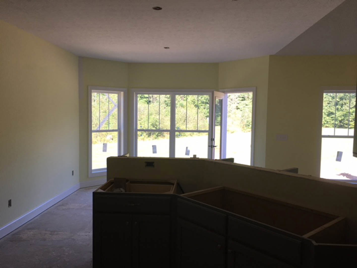 Modern kitchen corner with light wood drawers, large window with metal bars, view of leafy trees outside, dark accent wall, and neutral flooring.