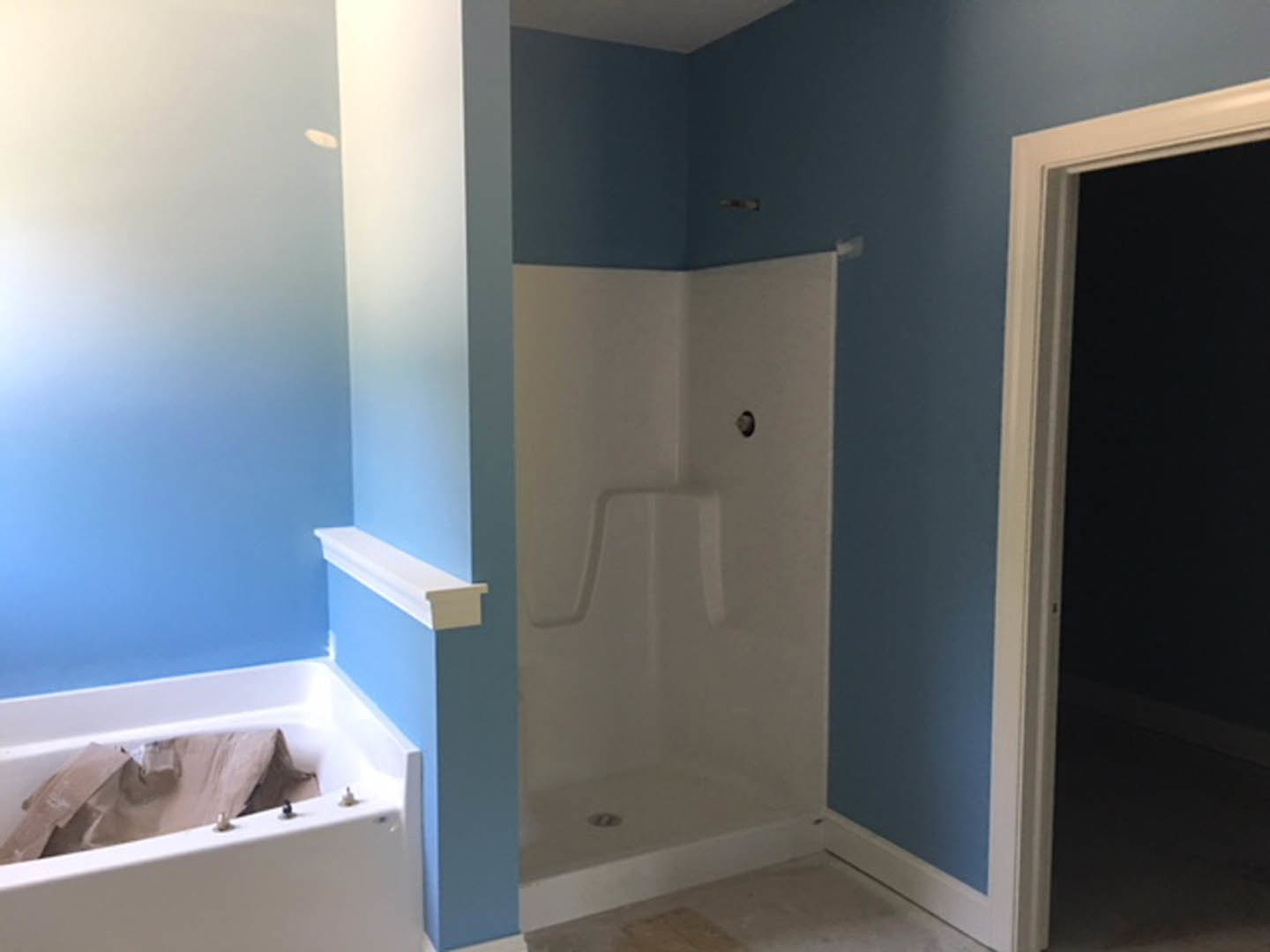 Freestanding white bathtub with a brown bag inside, glass shower enclosure with white frame, blue accent wall with white shelf, light-colored tile flooring, white door with