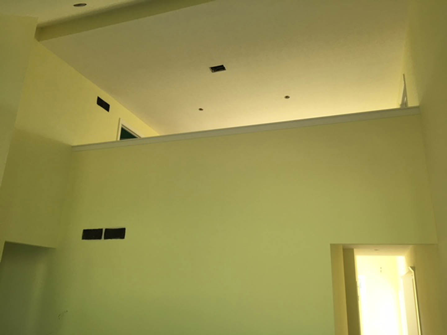 White ceiling with a central square light fixture, green accent wall featuring black square details, doorway allowing natural light, yellow wall visible in foreground.