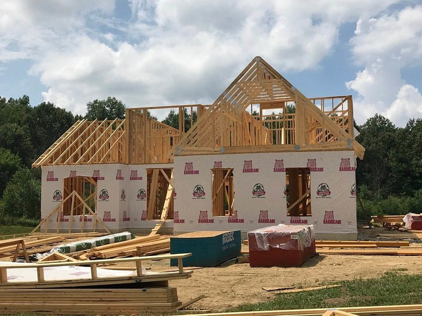 Wood-framed house under construction with exposed beams, plastic-wrapped box, large green dumpster with white text, pile of lumber, partially finished roof, and trees in the