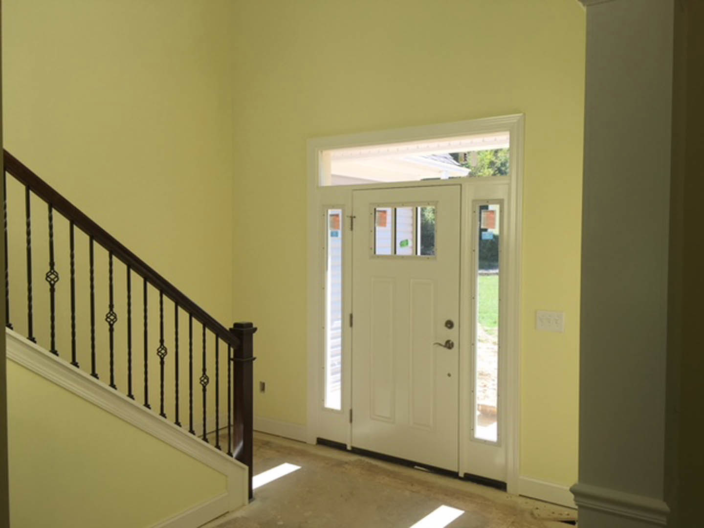 White paneled door with glass window, adjacent yellow wall with white electrical outlet, staircase with wooden handrail and white balusters, hardwood flooring
