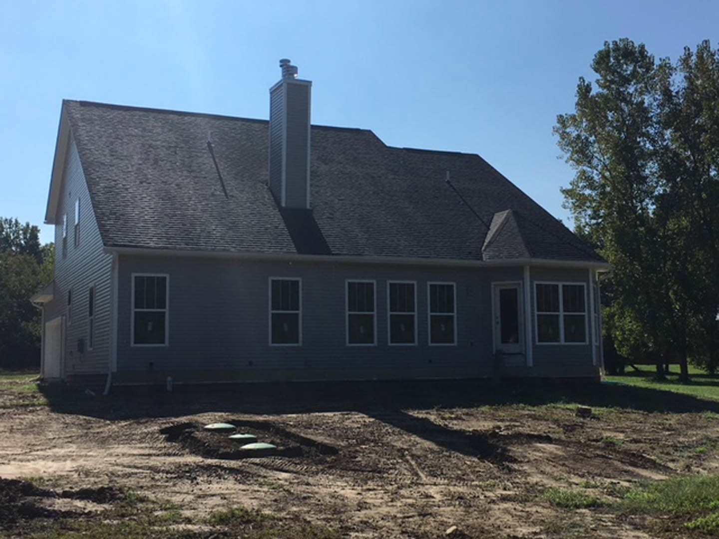 Two-story farmhouse with white-framed windows, brick chimney, and dirt yard featuring a blue pipe; leafy tree stands beside the house under a clear sky.