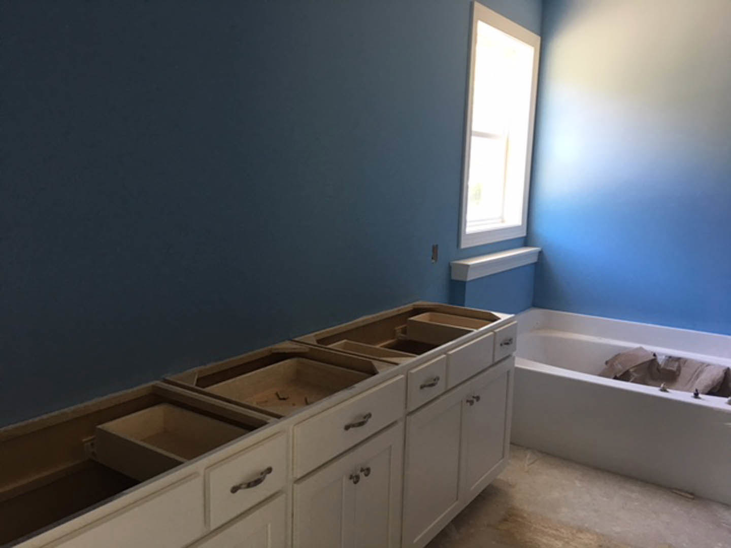 White bathroom featuring shaker-style cabinets, freestanding bathtub beneath a window, chrome fixtures, and light streaming onto tile flooring.