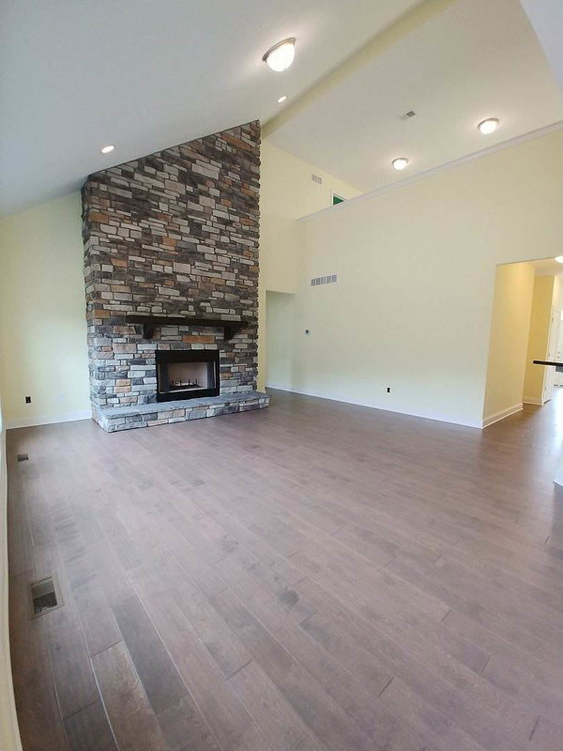 Spacious living room featuring a stone fireplace with glass window and wood mantel, hardwood flooring, plaster walls, ceiling light fixture, and wall vent
