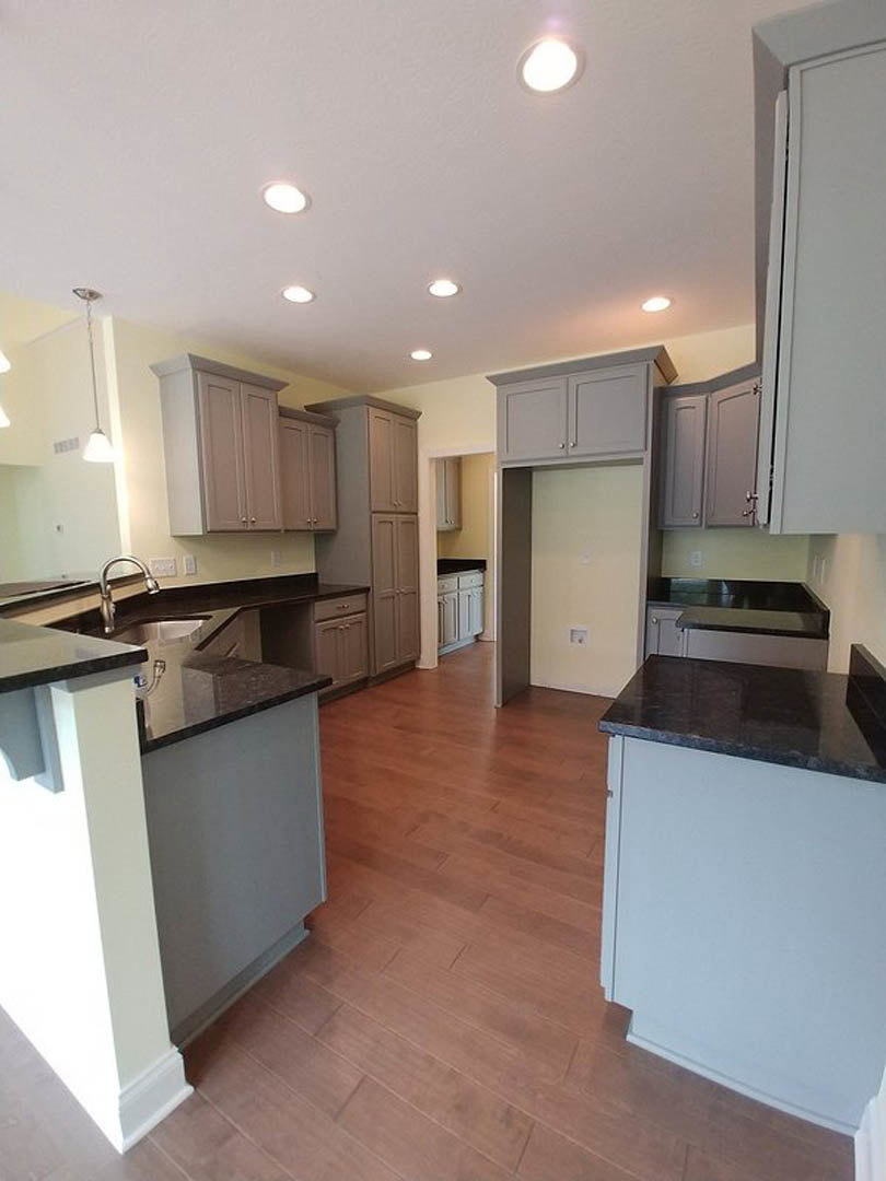 Grey shaker cabinets and matte black countertops in a kitchen with wood flooring, recessed ceiling light, stainless steel faucet, and white backsplash