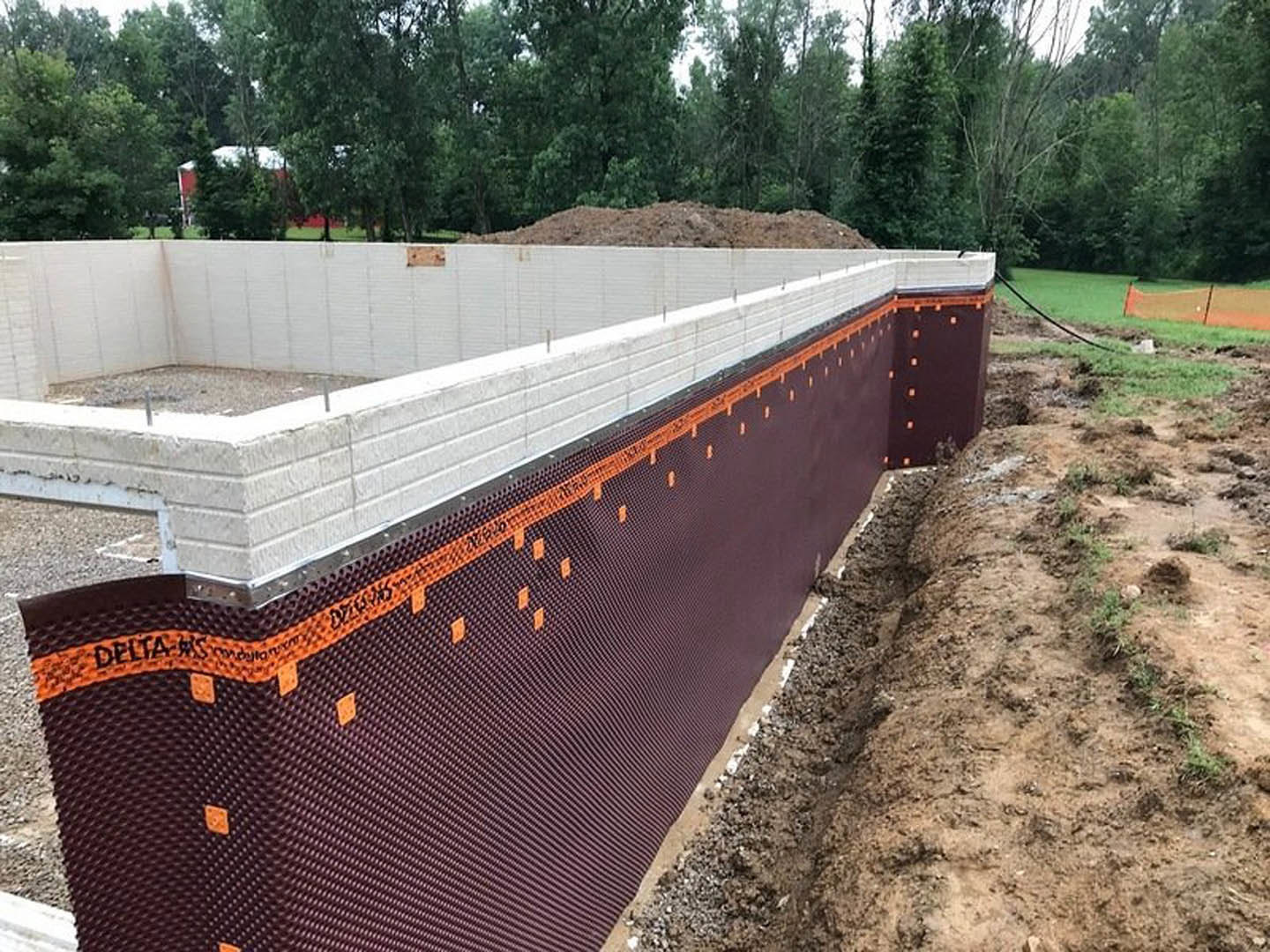 Brick foundation wall under construction with orange tape, surrounded by dirt, grass, and trees in the background