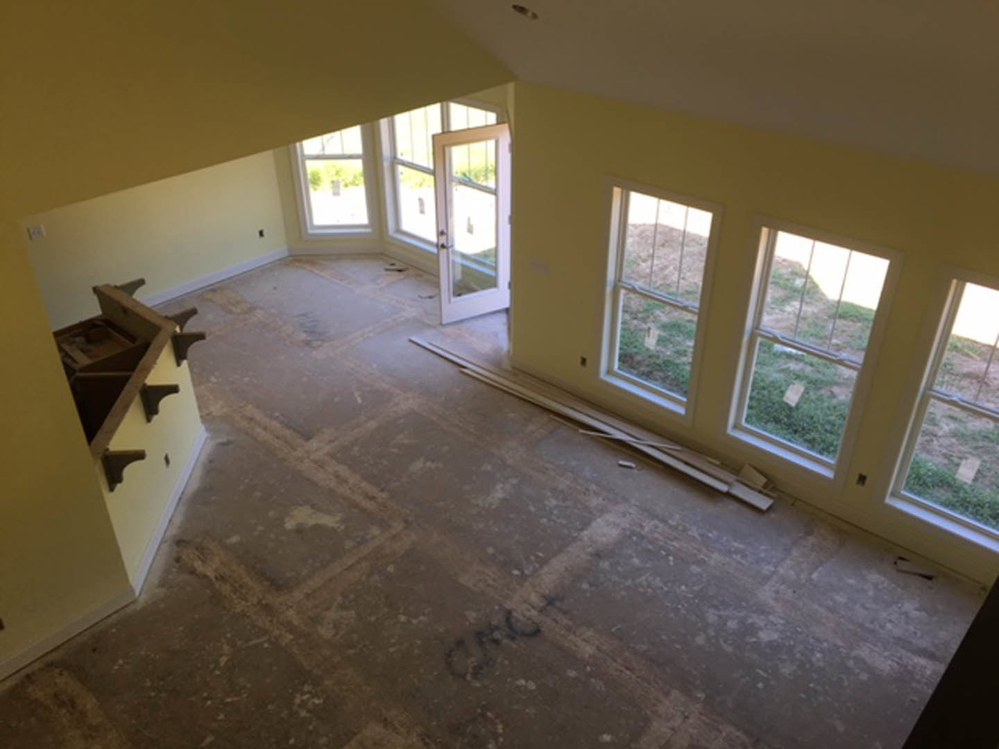 Bright room with light wood flooring, white plaster walls, multiple rectangular windows, and a single-pane door; built-in shelving visible along one wall.