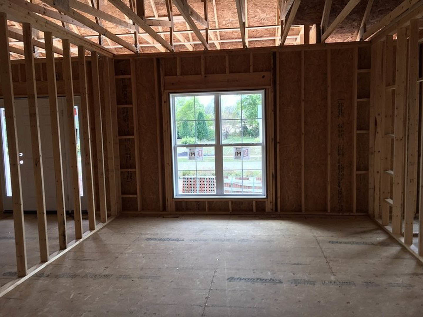 Room under construction with exposed wooden beams, window framed in wood, unfinished flooring with visible hole, and view of trees outside