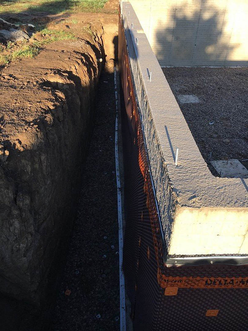 Concrete wall with a circular hole in the ground, stick resting on the surface, tree shadow cast across the wall, patch of grass and rocks nearby.