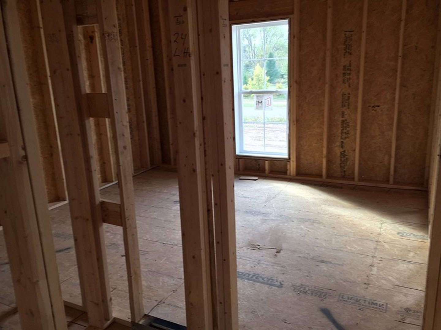 Room under construction with exposed wood framing, large window showing outdoor sign, wall featuring black text, wooden beam overhead, book resting on floor, bat-shaped shadow cast