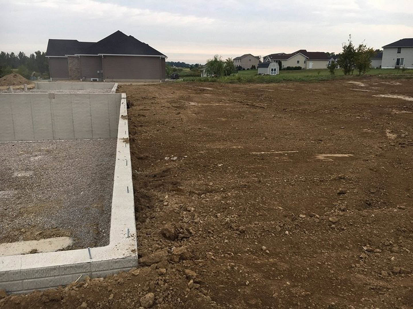 Concrete foundation and partially built exterior walls on a dirt lot, roof framing visible, surrounded by grass, trees, and neighboring buildings under a blue sky with scattered