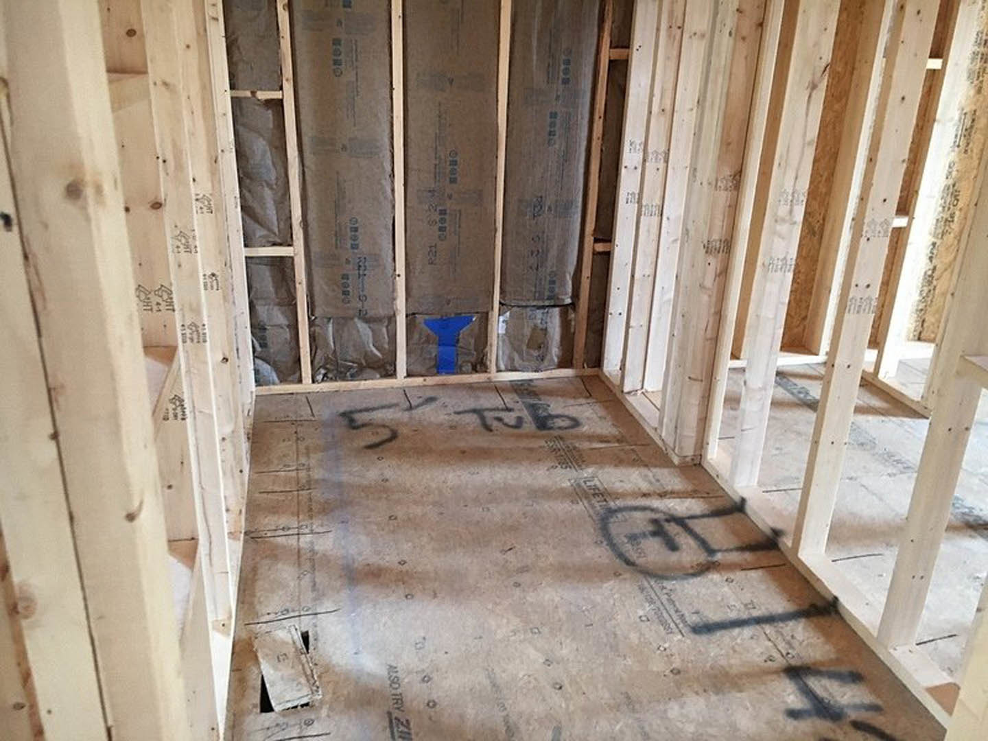 Room under construction with exposed wood framing, plywood subfloor marked with handwritten notes, and a blue handled tool resting on the floor