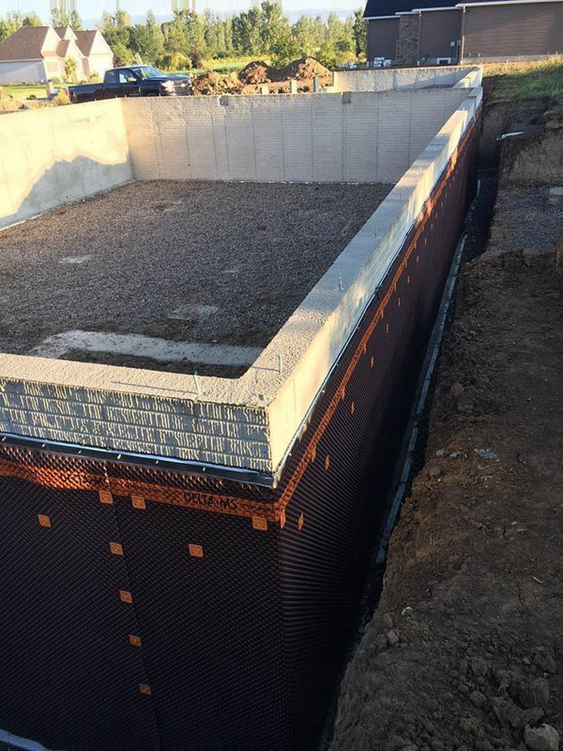 Concrete foundation slab surrounded by dirt, with a few trees nearby and a white-roofed building in the background.