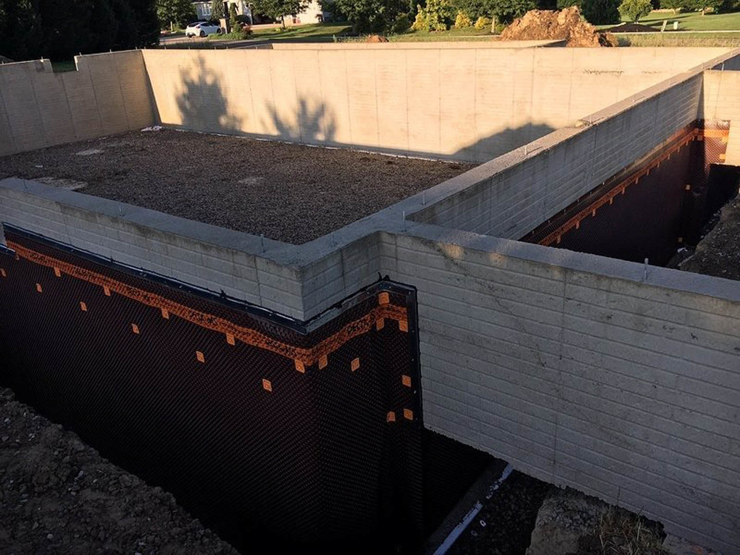 Concrete foundation wall with a circular hole, black and orange waterproofing membrane, brown and black surface, grassy area and trees in background.