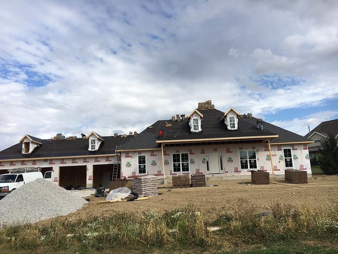Partially built house with exposed roof framing, scattered bricks and gravel in foreground, construction truck parked nearby, grassy yard, cloudy sky overhead