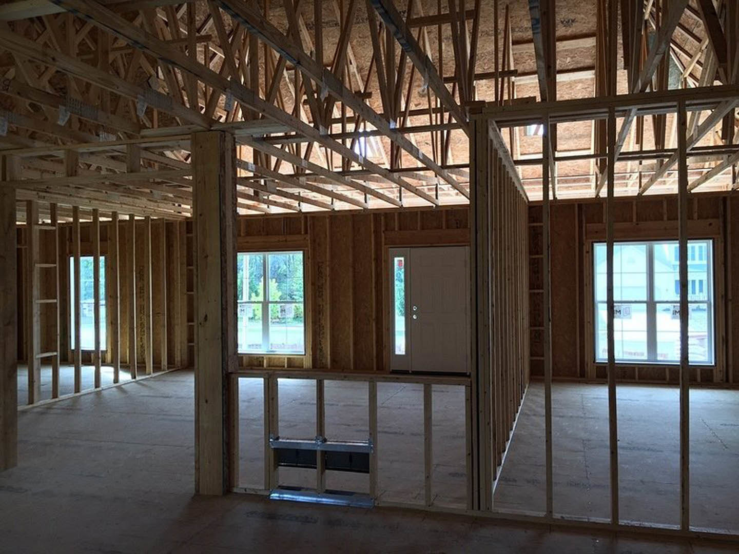 Wood-framed room under construction with exposed beams, unfinished floor, door opening, and multiple windows allowing daylight