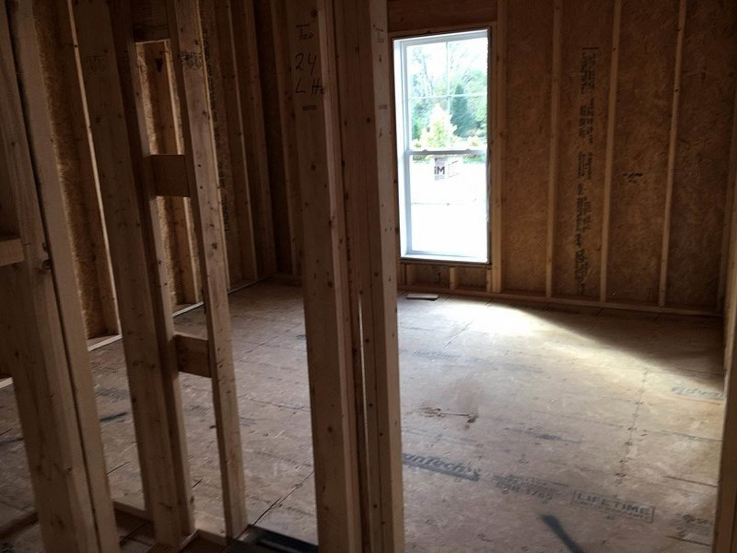 Room under construction with exposed wood framing, large window overlooking a tree, unfinished flooring illuminated by natural light, and wall marked with black text.