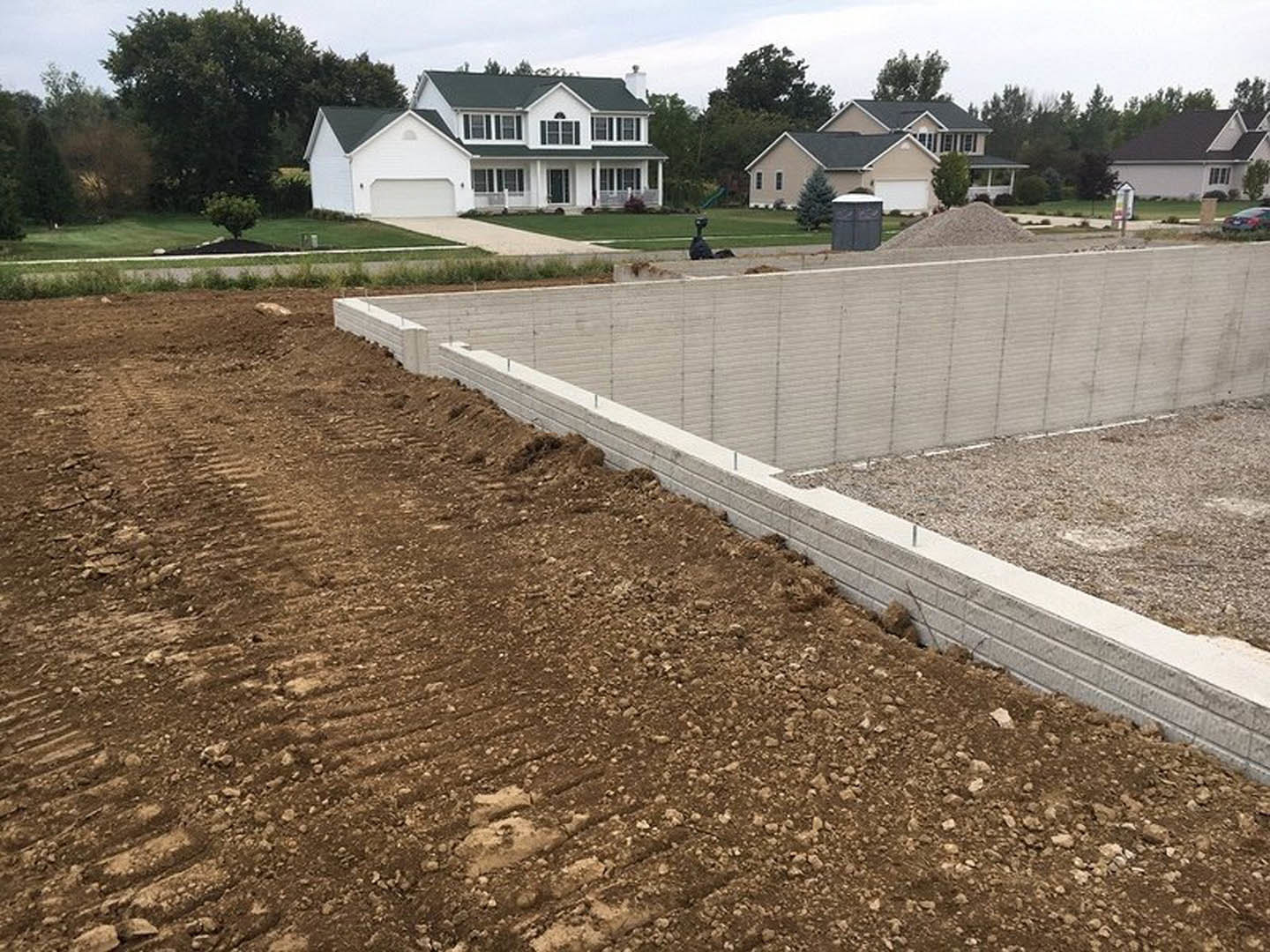Modern house with green roof, grey concrete foundation, white trim, fenced concrete retaining wall, grassy yard with tree, blue sky overhead