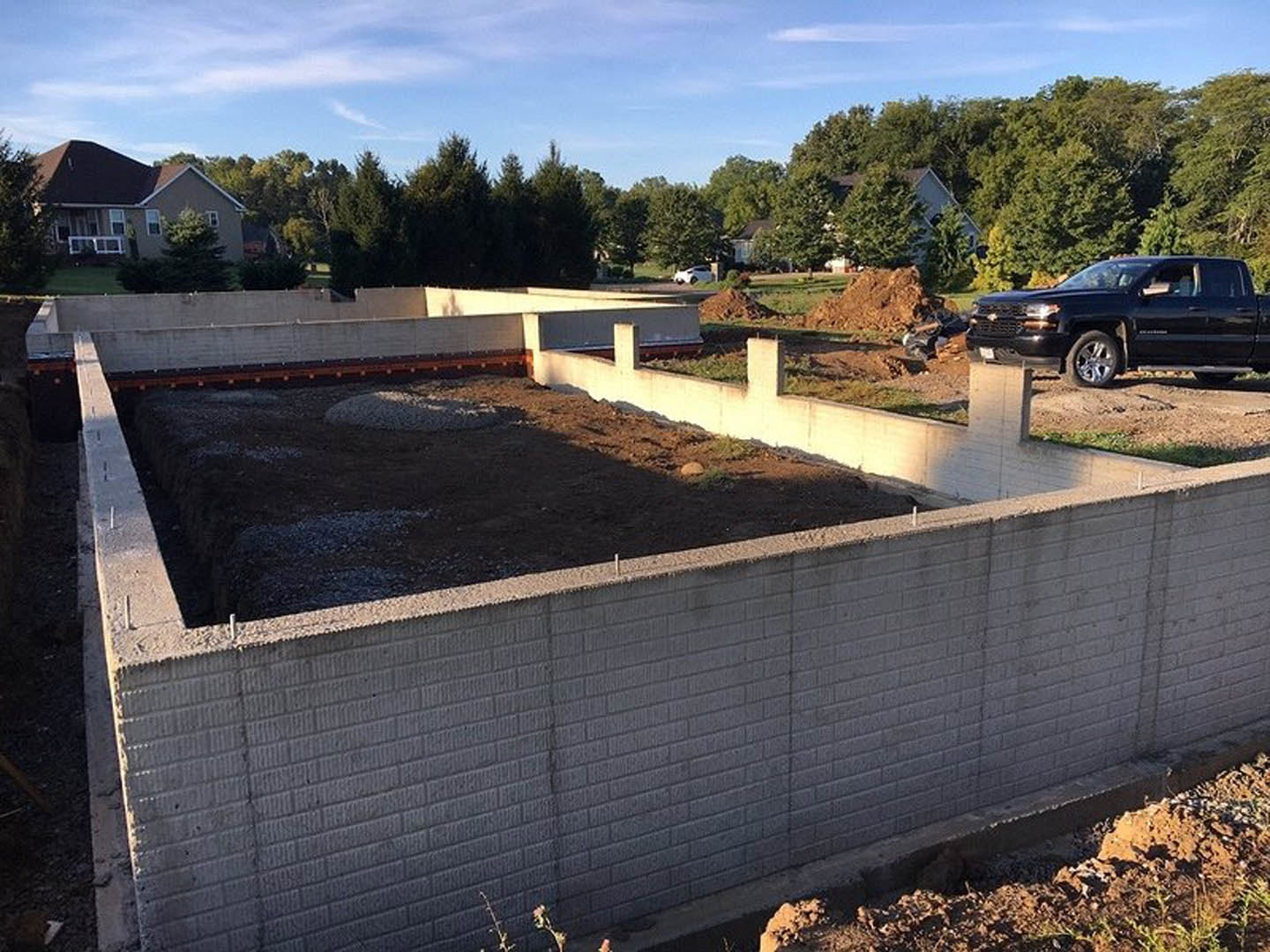 Black pickup truck parked beside unfinished concrete foundation, house structure in background, surrounded by trees under partly cloudy blue sky.