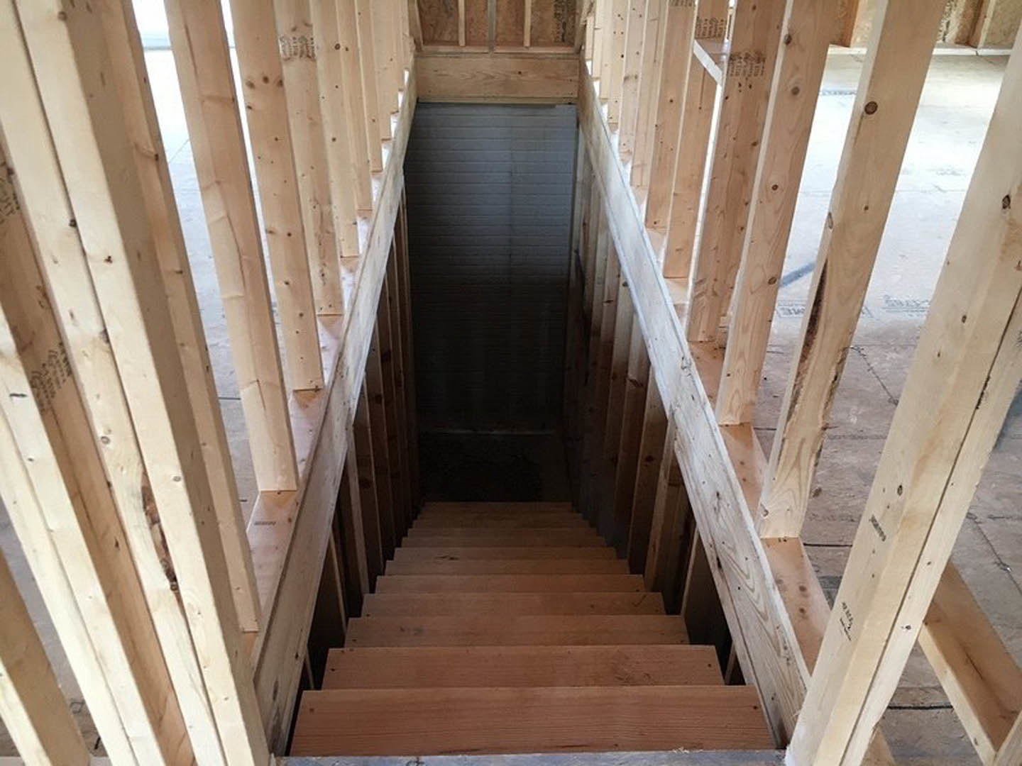 Wooden staircase with matching railing ascending from a dimly lit basement, light fixture at the top, door visible in the background, natural wood planks and surfaces throughout.