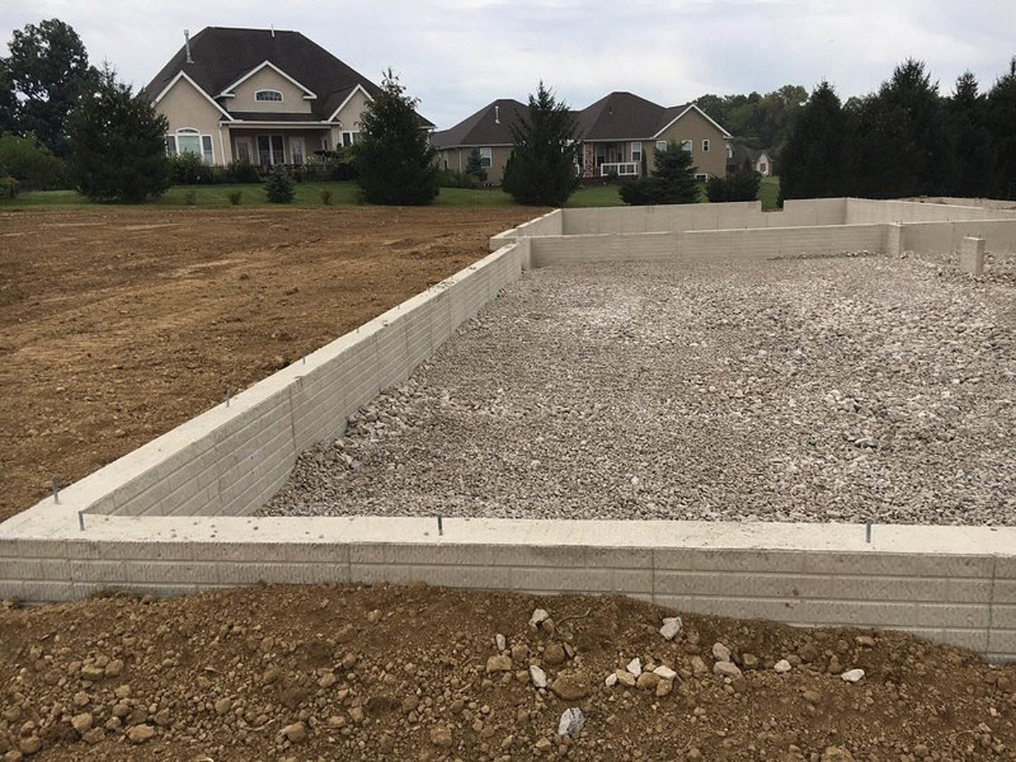 Concrete foundation with gravel and dirt, partially constructed brick wall, black roof, grassy lot with trees, cloudy sky in background
