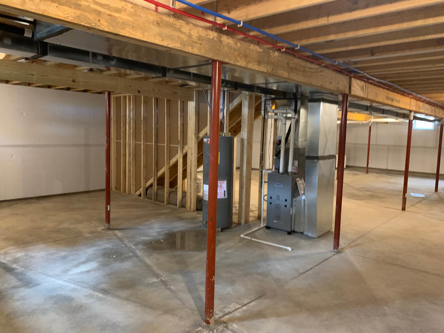 Basement room with exposed steel beams, red and blue plumbing pipes, wooden framing, white insulated panels, and a water tank.