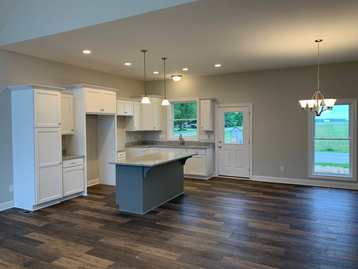 Kitchen with wood flooring, central island featuring granite countertops, blue base cabinetry, white door with window, large window overlooking a field, ceiling-mounted light