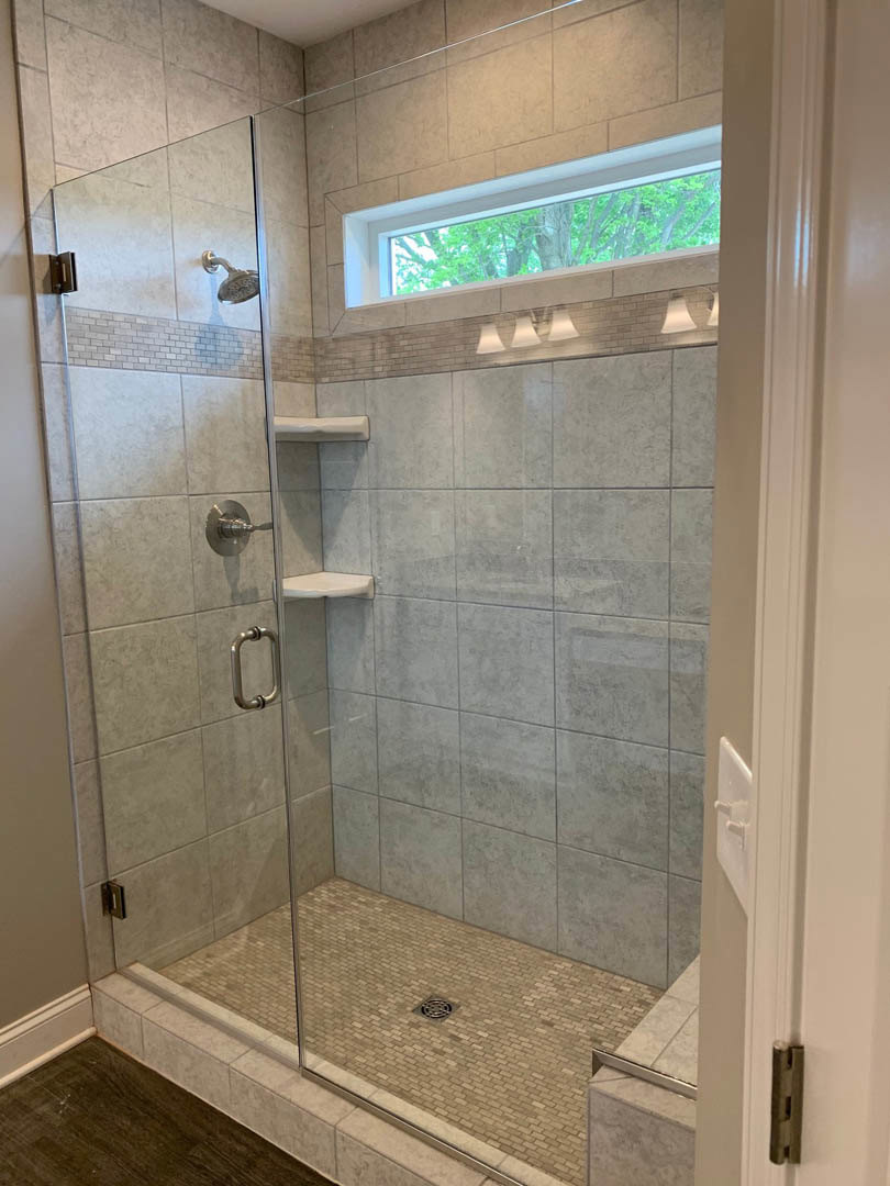 Walk-in shower with clear glass door, large round shower head, tiled walls and floor, metal handle, window overlooking trees, linear drain, adjacent wood flooring