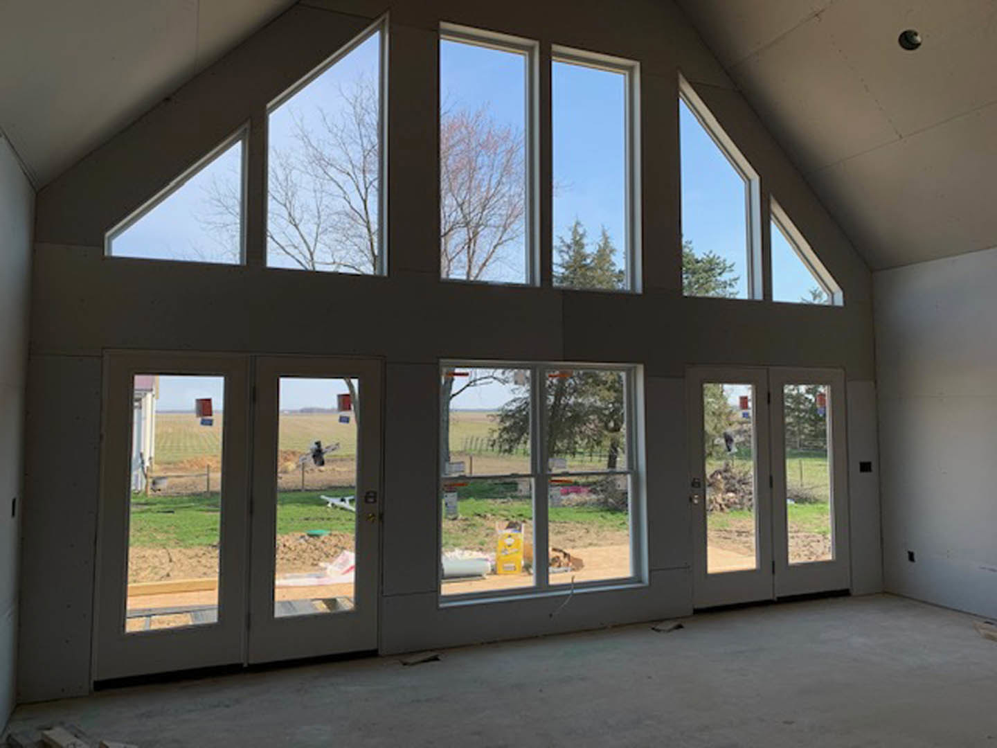 Spacious living area with multiple tall windows, light wood flooring, glass double doors, and outdoor views of trees and fields