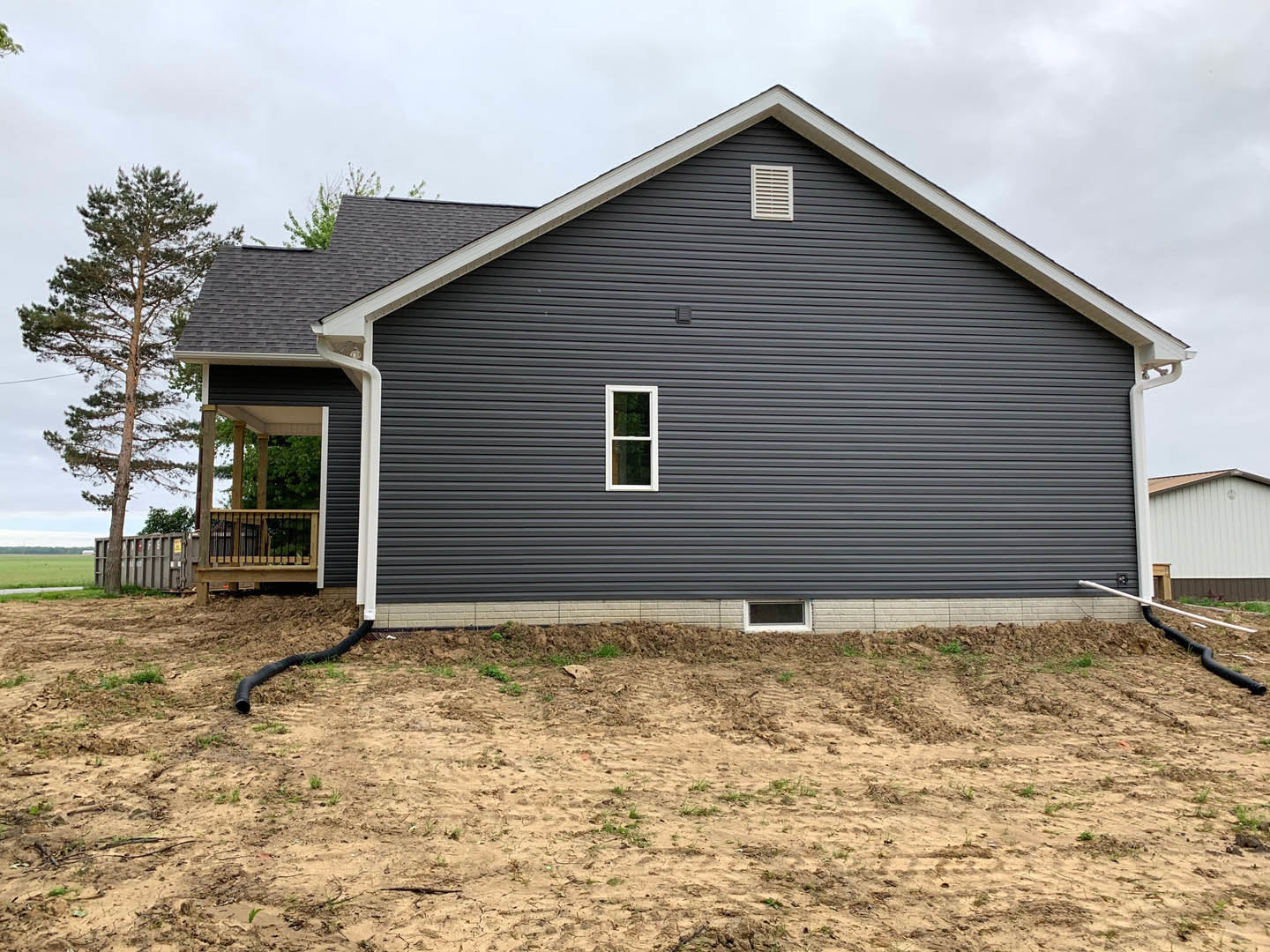 Two-story house with grey siding, white-framed windows, elevated wooden deck, white vent, fenced backyard, tall tree, and patchy grass over dirt lot under partly cloudy sky