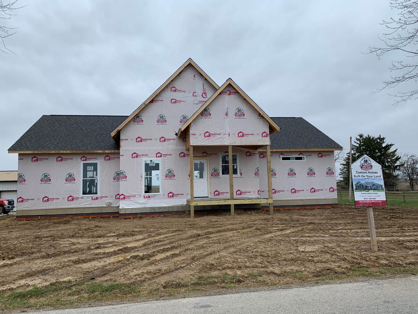 Two-story house under construction with plastic sheeting covering exterior walls, dirt lot with tire tracks in foreground, construction signs featuring house images mounted on