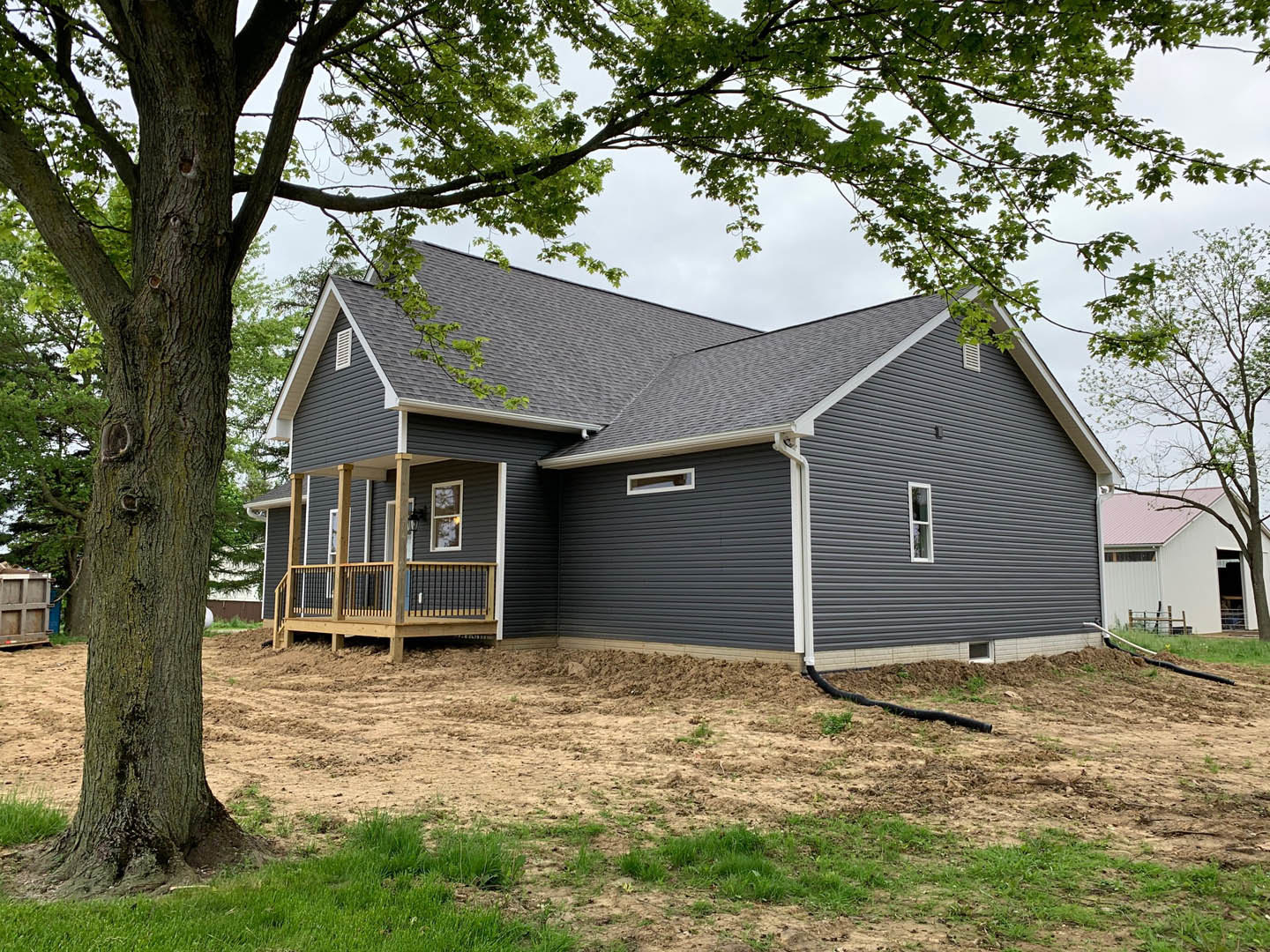 Two-story cottage with white siding and a covered porch, wooden deck with railing, large mature tree in front yard, patchy grass and dirt, small garden shed visible in background.