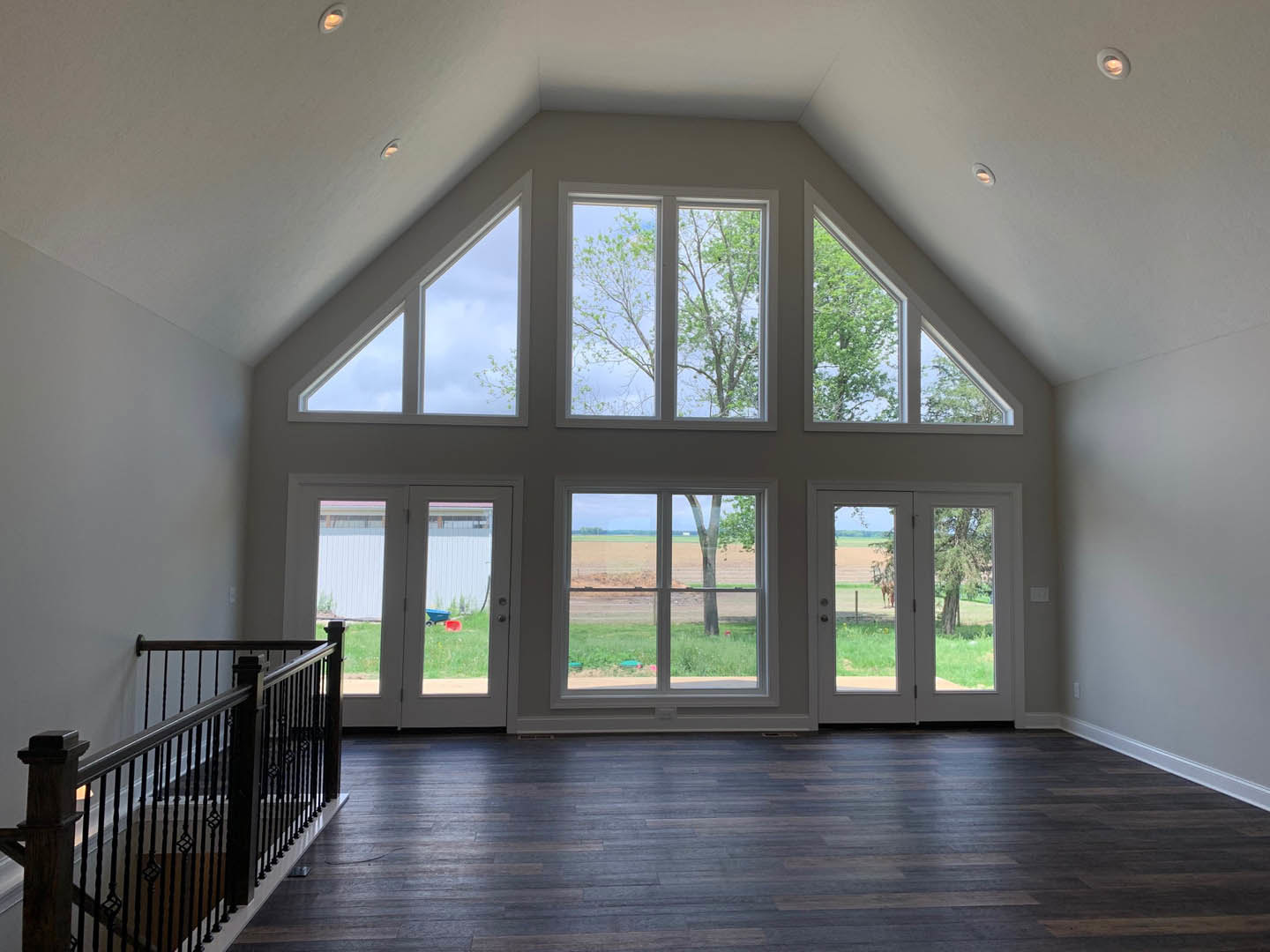 Spacious interior with hardwood flooring, black staircase railing, large white walls, multiple windows overlooking a field and trees, and a balcony area