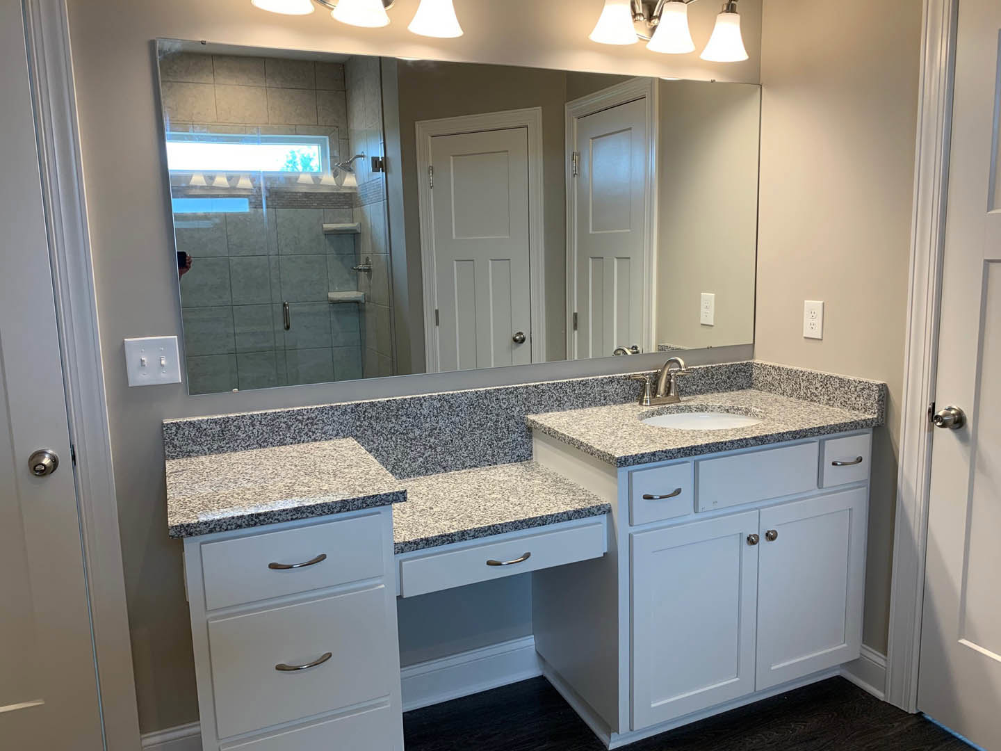 White bathroom vanity with silver drawer handles, quartz countertop, rectangular mirror above sink, white door with chrome handle, light switch on tiled wall.