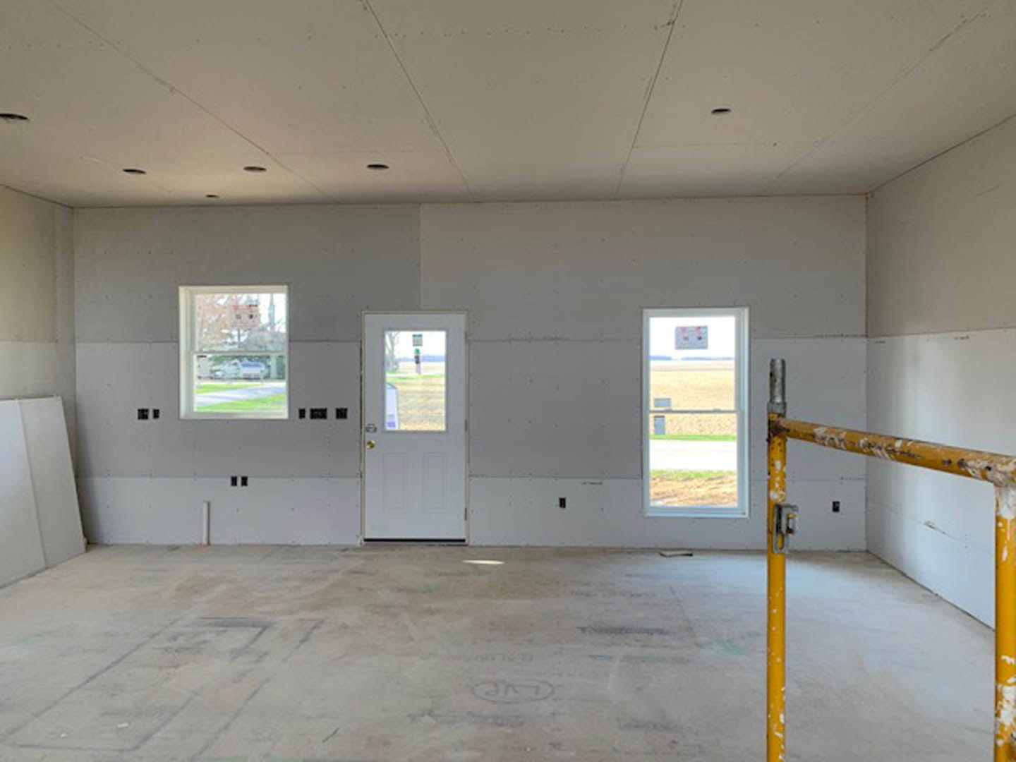 Bright room with white plaster walls, concrete floor, white door with glass window, large window overlooking green lawn and open field, yellow pipe along wall, unfinished ceiling.