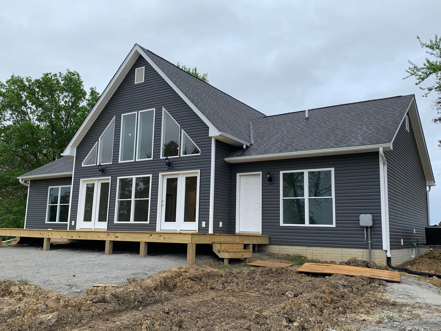 Two-story home under construction with white siding, glass panel door, white-framed windows, covered porch, wooden deck, dirt yard, and surrounding trees
